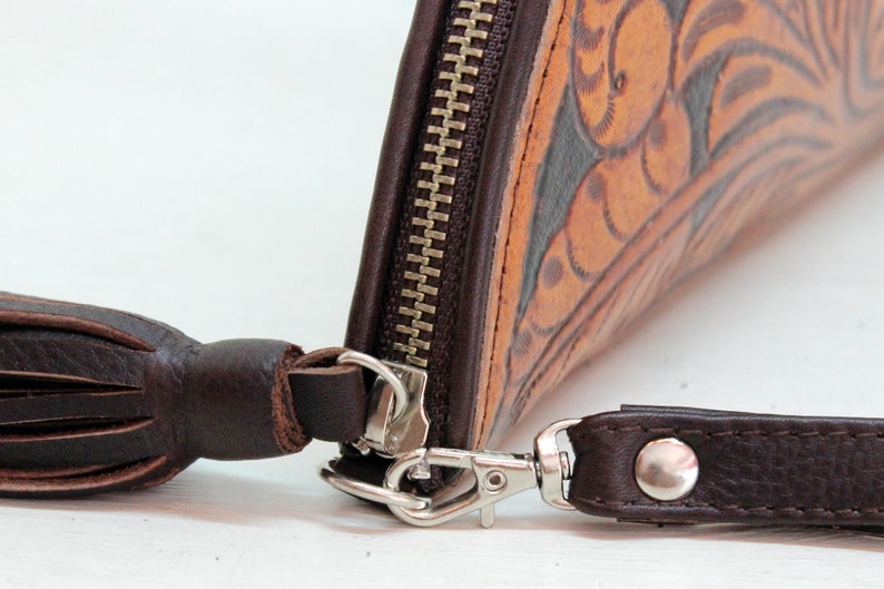 May include: A brown leather wallet with a zipper closure and a tassel. The wallet has a floral design and a silver clasp.