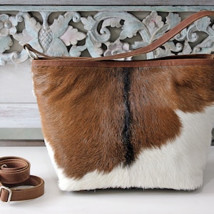 May include: A brown and white cowhide tote bag with a brown leather strap. The bag is sitting on a white surface with a carved wooden background.
