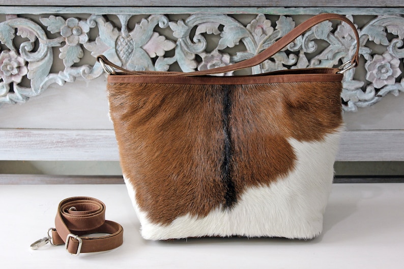 May include: A brown and white cowhide tote bag with a brown leather strap. The bag is sitting on a white surface with a carved wooden background.