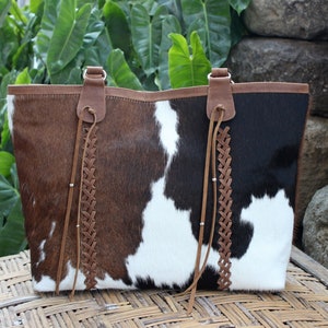 May include: A brown and black cowhide tote bag with braided leather straps and tassels. The bag has a white and brown patchwork design.