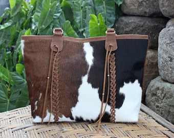 Puede incluir: Un bolso tote de piel de vaca marrón y negro con correas de cuero trenzadas y borlas. El bolso tiene un diseño de patchwork blanco y marrón.