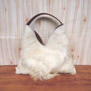 May include: White fur handbag with a brown leather handle. The bag has a unique, triangular shape.
