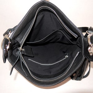May include: Black leather shoulder bag with a silver zip and a silver ring on the strap. The bag has a flap closure and a small pocket on the inside.