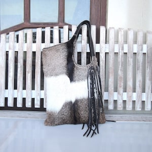 May include: A large tote bag made of cowhide with a patchwork design of gray, white, and black fur. The bag has a black leather handle and decorative fringe.