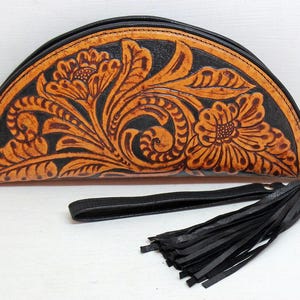 May include: A half-moon shaped leather clutch with a black tassel. The clutch is decorated with a floral pattern in shades of brown and orange.