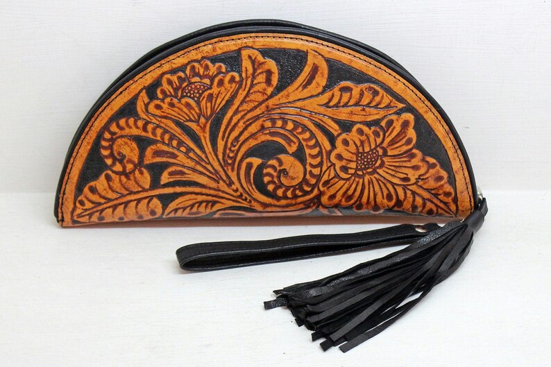 May include: A half-moon shaped leather clutch with a black tassel. The clutch is decorated with a floral pattern in shades of brown and orange.