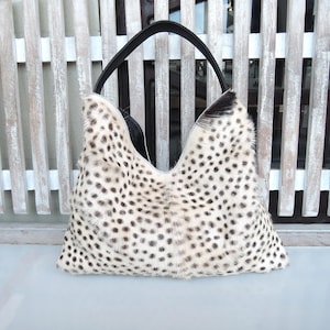 May include: A hobo-style handbag featuring a cream-colored exterior with a dense pattern of dark brown and black spots. The bag has a single, smooth black shoulder strap and a V-shaped top opening, showcasing a textured, fur-like material.