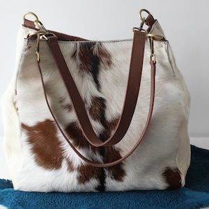 Puede incluir: Un bolso tote de piel de vaca blanca y marr&oacute;n con correas de cuero marr&oacute;n y herrajes dorados.