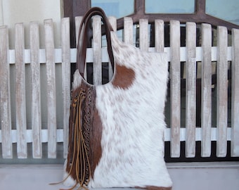 May include: A tote bag crafted from white and brown cowhide with a textured hair-on-hide finish. The bag features a dark brown leather handle, a vertical panel of dark brown leather with decorative lacing, and long leather fringe tassels.