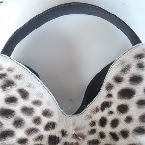 Puede incluir: Un bolso de estilo hobo con un patr&oacute;n de estampado animal en blanco y negro y un acabado texturizado de pelo. El bolso tiene una abertura profunda en forma de V en la parte superior y un asa de cuero negro liso que se arquea sobre el bolso.