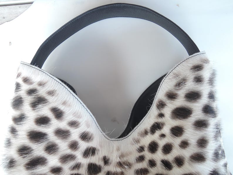 Puede incluir: Un bolso de estilo hobo con un patr&oacute;n de estampado animal en blanco y negro y un acabado texturizado de pelo. El bolso tiene una abertura profunda en forma de V en la parte superior y un asa de cuero negro liso que se arquea sobre el bolso.