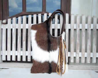 May include: A shoulder bag crafted from textured cowhide featuring a tri-colour pattern of brown, white, and black fur. The bag includes a dark brown leather handle and decorative leather fringe tassels hanging from the side with metal hardware accents.