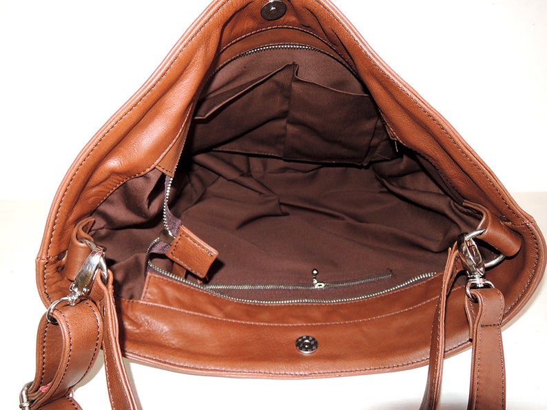 May include: A brown leather shoulder bag with a zipper closure and a flap. The bag has a brown lining and a small pocket inside. The bag has a long adjustable strap.