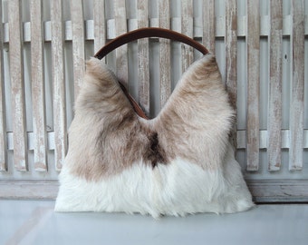 May include: A hobo-style handbag crafted from textured hair-on hide featuring a color-blocked pattern of white and light brown fur with a dark brown patch. The bag includes a smooth, solid brown leather shoulder strap attached to the top opening.