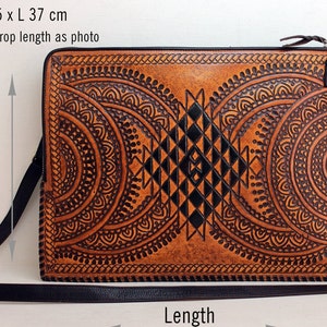 Puede incluir: Una funda para portátil de cuero marrón con un diseño geométrico negro. La funda tiene una correa ajustable negra y un cierre de cremallera. Las dimensiones son 26,5 cm de alto por 37 cm de largo.