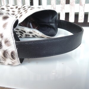 Puede incluir: Un bolso de estilo hobo con un exterior de piel con estampado animal en blanco y negro. El bolso incluye una correa de hombro ancha de cuero negro texturizado y un forro interior oscuro visible a trav&eacute;s de la parte superior abierta. La construcci&oacute;n muestra costuras limpias.