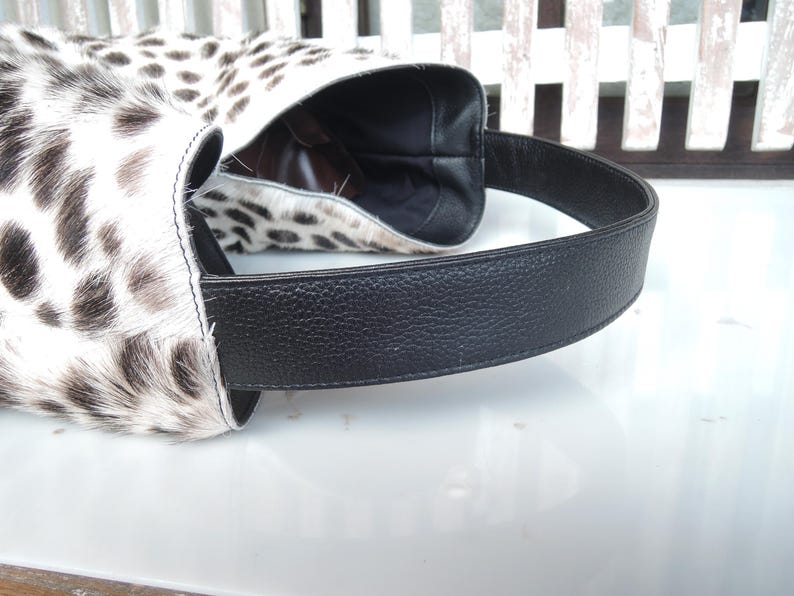 Puede incluir: Un bolso de estilo hobo con un exterior de piel con estampado animal en blanco y negro. El bolso incluye una correa de hombro ancha de cuero negro texturizado y un forro interior oscuro visible a trav&eacute;s de la parte superior abierta. La construcci&oacute;n muestra costuras limpias.