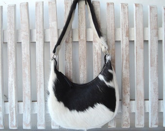 May include: A crescent-shaped shoulder bag crafted from cowhide featuring a distinct black and white spotted pattern. The bag includes a single matching fur strap attached with silver-toned metal hardware and a top zipper closure for access.