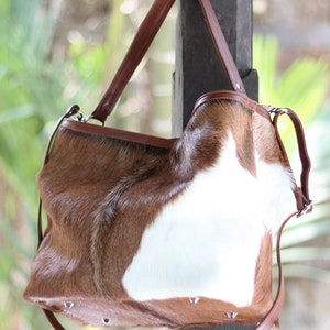 May include: A brown and white cowhide handbag with a brown leather strap. The bag has a flap closure and is decorated with silver studs.