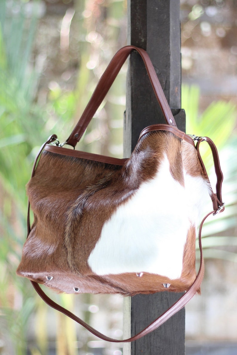 May include: A brown and white cowhide handbag with a brown leather strap. The bag has a flap closure and is decorated with silver studs.