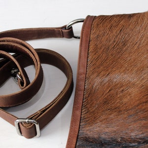 May include: A brown leather crossbody bag with a white and brown cowhide pattern. The bag has a long adjustable strap and a silver buckle.