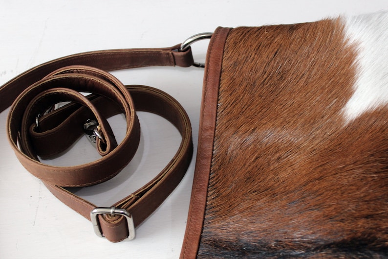 May include: A brown leather crossbody bag with a white and brown cowhide pattern. The bag has a long adjustable strap and a silver buckle.