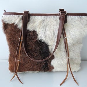 May include: A cowhide tote bag with a patchwork design in shades of brown, black, and white. The bag features brown leather handles and trim, with decorative leather tassels hanging from the sides. The bag is designed for carrying items.