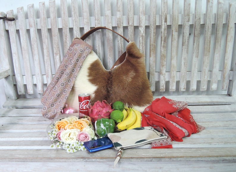 Puede incluir: Un bolso hobo de piel de vaca marr&oacute;n y blanco con correa de cuero, acompa&ntilde;ado de una lata roja de Coca-Cola, una fruta del drag&oacute;n, limas verdes, un pimiento verde, pl&aacute;tanos amarillos, un ramo de rosas, un smartphone azul y una bufanda roja.