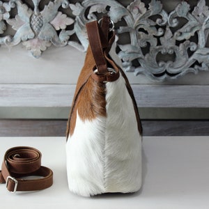 May include: A brown and white cowhide tote bag with a brown leather strap. The bag has a top handle and a detachable shoulder strap.