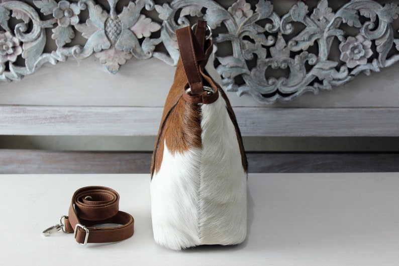 May include: A brown and white cowhide tote bag with a brown leather strap. The bag has a top handle and a detachable shoulder strap.