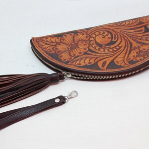 May include: A brown leather half-moon clutch with a floral tooled design. The clutch has a zipper closure and a brown leather tassel and strap.