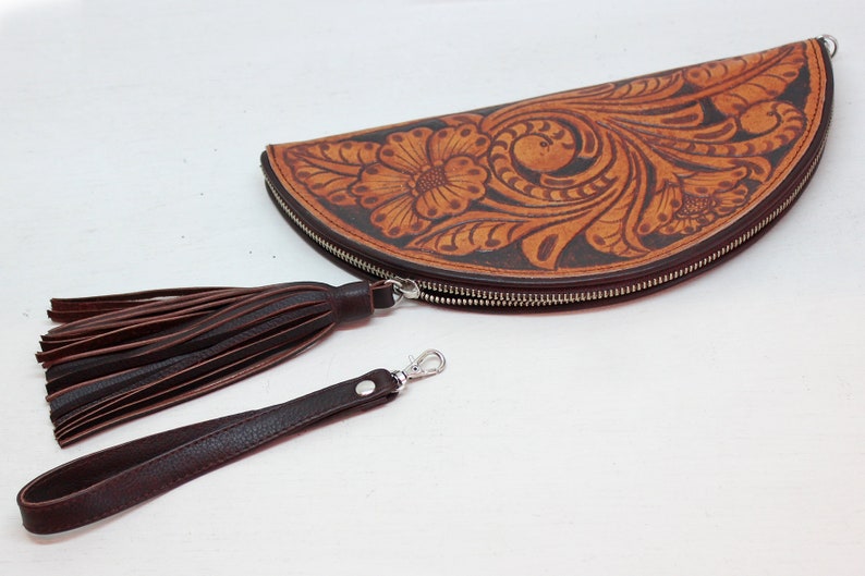 May include: A brown leather half-moon clutch with a floral tooled design. The clutch has a zipper closure and a brown leather tassel and strap.