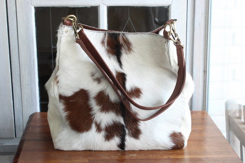 Puede incluir: Un bolso tote de piel de vaca blanca y marr&oacute;n con correas de cuero marr&oacute;n y herrajes dorados.