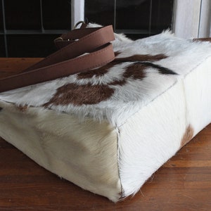 Puede incluir: Un bolso tote de piel de vaca blanca y marr&oacute;n con correas de cuero marr&oacute;n. El bolso tiene una forma rectangular y est&aacute; hecho de un material suave y natural.