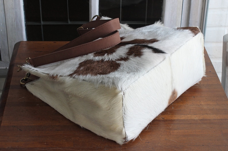 Puede incluir: Un bolso tote de piel de vaca blanca y marr&oacute;n con correas de cuero marr&oacute;n. El bolso tiene una forma rectangular y est&aacute; hecho de un material suave y natural.