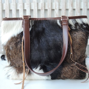 May include: A brown, black, and white cowhide tote bag with brown leather straps and tassels.