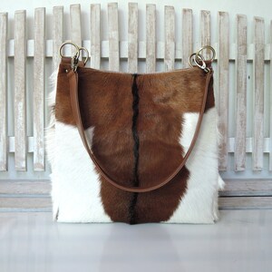 May include: A rectangular tote bag with a brown and white cowhide pattern. The bag has a brown leather strap and gold-colored metal rings. The bag is set against a white wooden fence.