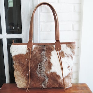 May include: A brown and white cowhide tote bag with leather straps and a brown leather trim. The bag has a large main compartment and a zipper closure.