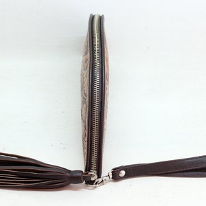May include: A brown leather clutch with a zipper closure and a tassel. The clutch has a decorative embossed pattern and a brown leather strap.