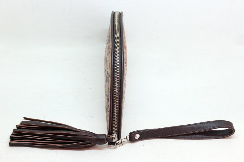 May include: A brown leather clutch with a zipper closure and a tassel. The clutch has a decorative embossed pattern and a brown leather strap.
