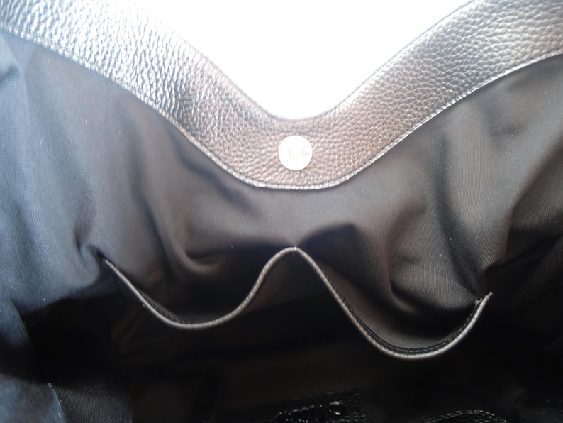 Puede incluir: El interior de un bolso negro con un borde de cuero texturizado en la parte superior y un cierre magn&eacute;tico plateado. El interior est&aacute; forrado con tela negra e incluye un bolsillo central abierto para la organizaci&oacute;n.