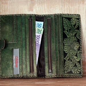 May include: A green leather wallet with a floral design and a zipper closure. The wallet is open and has several compartments for cash and cards. The wallet has a small label that says "Olive Culture & Craft".