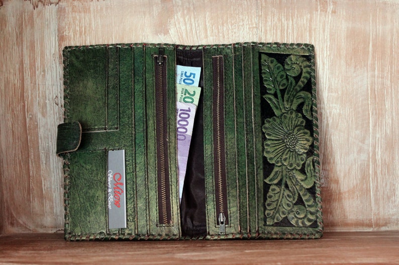 May include: A green leather wallet with a floral design and a zipper closure. The wallet is open and has several compartments for cash and cards. The wallet has a small label that says "Olive Culture & Craft".