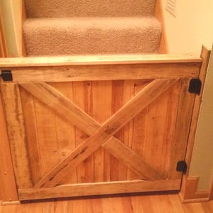 May include: A light brown wooden safety gate featuring a classic X-brace design across the front panel. The gate is constructed from vertical wooden planks and is secured to the wall and door frame with black metal hinges and a latch mechanism.