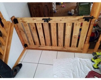 May include: A wooden baby gate with vertical slats and a latch. The gate is made of light-colored wood and has black hinges. It is designed to be placed in a doorway or opening to prevent a child from entering.