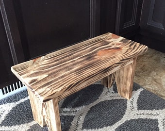 May include: A small wooden stool with a rustic, burnt finish. The stool has a rectangular top and two legs on each side. The stool is sitting on a gray and white patterned rug.