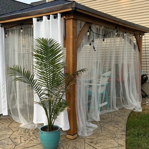 May include: A wooden gazebo with white sheer curtains and a potted palm tree. The gazebo has a brown wooden frame and a metal roof. There are fairy lights hanging from the roof.