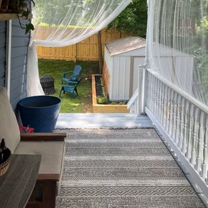 May include: A view from a porch with a white railing and a white sheer curtain. The porch has a gray and white patterned rug. A blue planter is on the porch floor. A wooden shed is visible in the backyard.