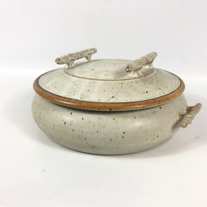 May include: A white ceramic serving dish with a lid and two handles. The dish has a speckled glaze and a brown rim. The lid has two wooden handles.