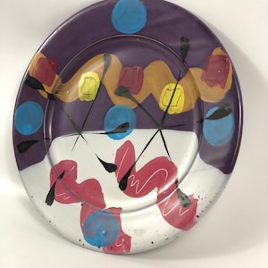 May include: A purple ceramic plate with a white center and abstract designs in black, red, yellow, and blue.